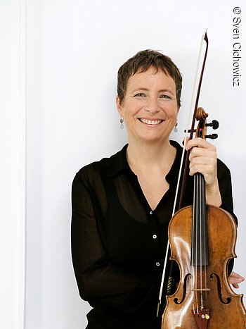 Christine Busch Meisterkonzert 2025 Eine schwarz gekleidete Dame mittleren Alters mit kurzen Haaren hält eine Violine in der Hand und lächelt.