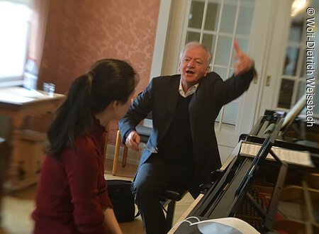 In einem mit Antiquitäten ausgestattetem Raum stehen zwei Flügel, an denen ein älterer Herr und eine junge Dame zusammen sitzen. Die Dame spielt, der Herr gestikuliert.