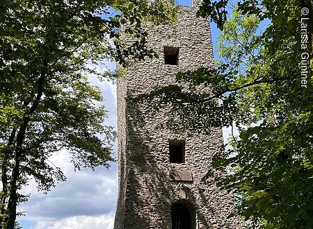 Steinerner Turm im Wald mit Bank im Vordergrund, blauer Himmel im Hintergrund.