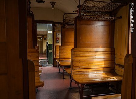 Innenraum eines historischen Zugwaggons mit Holzbänken und Gepäcknetzen.