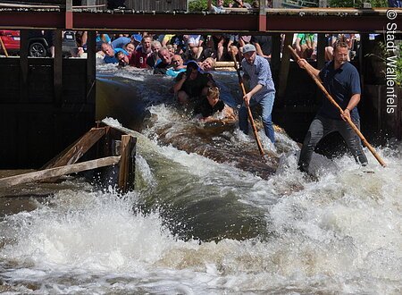 Gruppe von Menschen auf einem Floß in einem Fluss, umgeben von spritzendem Wasser.