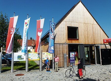 Modernes Gebäude mit Holzverkleidung, davor mehrere Fahnen und Informationstafeln. Eine Person mit Fahrrad steht vor dem Eingang.