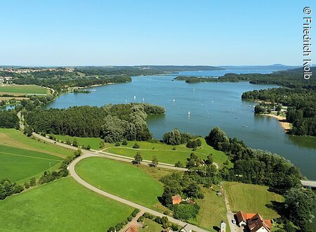 Luftaufnahme eines Sees mit Segelbooten, umgeben von Wäldern und Wiesen. Im Hintergrund sind Hügel sichtbar.