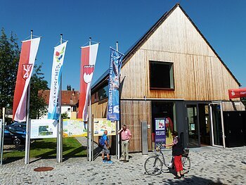 Modernes Gebäude mit Holzverkleidung, davor mehrere Fahnen und Informationstafeln. Eine Person mit Fahrrad steht vor dem Eingang.