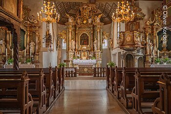 Innenraum einer barocken Kirche mit reich verziertem Altar, Holzbänken und Kronleuchtern.