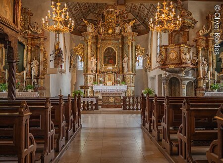 Innenraum einer barocken Kirche mit reich verziertem Altar, Holzbänken und Kronleuchtern.