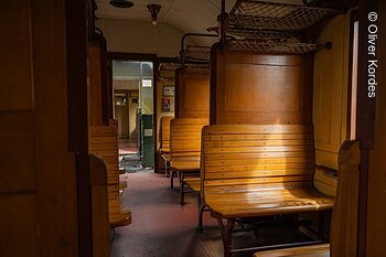 Innenraum eines historischen Zugwaggons mit Holzbänken und Gepäcknetzen.