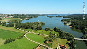Luftaufnahme eines Sees mit Segelbooten, umgeben von Wäldern und Wiesen. Im Hintergrund sind Hügel sichtbar.