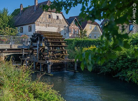 Altes Wasserrad an einem Fluss, umgeben von traditionellen Fachwerkhäusern und üppigem Grün.