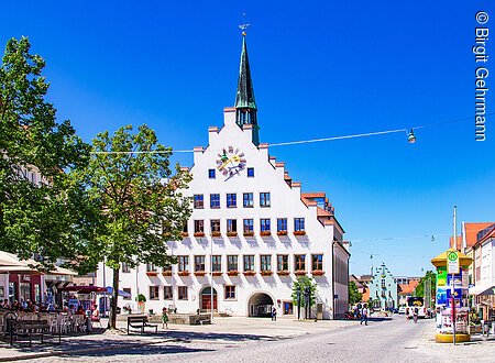 Ein historisches Gebäude mit Giebel und Turm steht auf einem Platz, umgeben von Bäumen und Cafés.