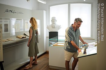 Zwei Personen betrachten Exponate in einem Museum, darunter Bücher und eine Büste.