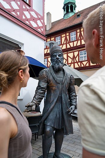 Zwei Personen betrachten eine Bronzestatue in historischer Kleidung vor Fachwerkhäusern.