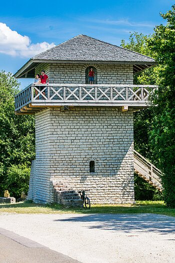 Steinturm mit Holzgeländer und Treppe, umgeben von Bäumen. Drei Personen auf dem Balkon. Fahrrad unten.