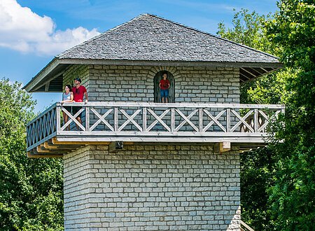 Steinturm mit Holzgeländer und Treppe, umgeben von Bäumen. Drei Personen auf dem Balkon. Fahrrad unten.