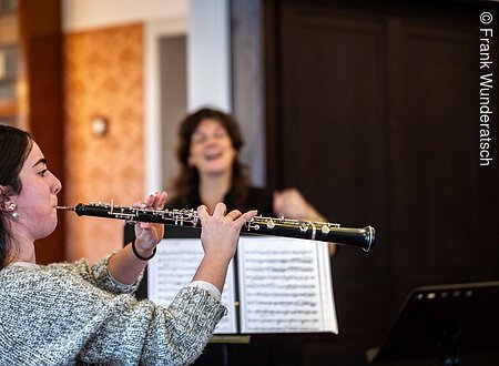 Eine junge Dame bläst in eine Oboe, neben ihr eine Dame, die mit ihr spricht und gestikuliert.