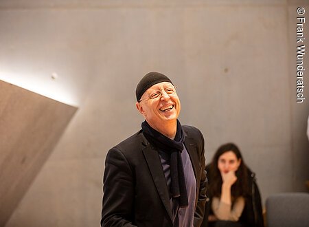 Ein Herr mit einer schwarzen Mütze steht vor einer Betonwand und lacht. Im Hintergrund ist unscharf eine sitzende junge Frau zu sehen.