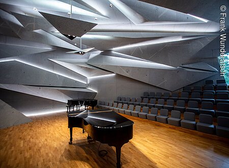 Zwei schwarze Konzertflügel mit Hocker stehen in einem mit Parkett ausgelegten großen Raum mit steinernen, spitz zulaufenden Bauelementen aus Stein, die geheimnisvoll beleuchtet und ineinander verschachtelt sind. Im Hintergrund sind Stuhlreihen zu erkennen.