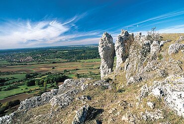 Felsformation auf einem Hügel mit Blick auf Felder, Wald und Dorf unter blauem Himmel mit Wolken.