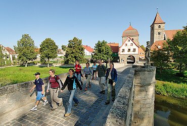 Gruppe von neun Personen wandert auf einer gepflasterten Brücke in einem Dorf mit historischen Gebäuden und Bäumen.