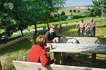 Zwei Frauen sitzen an einem Holztisch im Freien, zwei Männer schieben Fahrräder auf einer Wiese im Hintergrund.