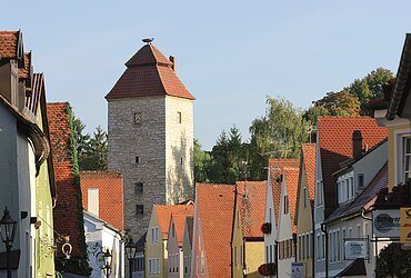 Straße mit bunten Häusern und einem hohen Steinturm mit rotem Dach im Hintergrund bei klarem Himmel.