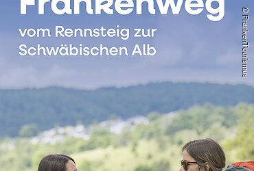 Zwei Frauen mit Rucksäcken sitzen auf einer Wiese und blicken in eine grüne Landschaft mit Hügeln.