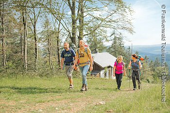 Vier Wanderer mit Rucksäcken gehen auf einem Waldweg an einem Haus vorbei bei Tageslicht.