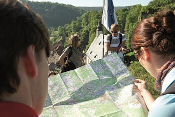 Vier Personen mit Rucksäcken schauen auf eine Wanderkarte vor einem Dorf mit Kirche und bewaldeten Hügeln.