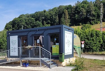 Kleines Verkaufsgebäude aus Metall mit Rampe, umgeben von Bäumen und Straße im Hintergrund.