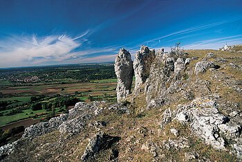 Felsformationen auf einem Hügel mit Blick auf Felder, Wald und Dorf unter blauem Himmel mit Wolken.