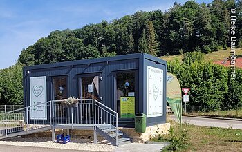 Kleines Verkaufsgebäude aus Metall mit Rampe, umgeben von Bäumen und Straße im Hintergrund.