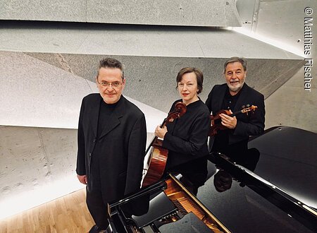 Eine Frau mit Violoncello und zwei Männer, einer davon mit Geige, stehen in einem Saal neben einem Flügel.