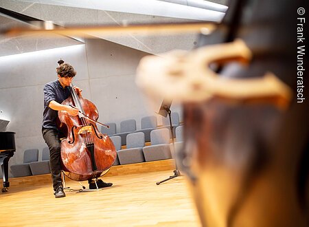 Kontrabass-Unterricht im Konzertsaal Eine Person mit einem Kontrabass steht mit seinem Instrument in einem Raum mit Gesteinselementen. Er setzt den Bogen an..Am, rechten Bildrand ist unscharf der Steg eiees weiteren Kontrabasses zu sehen, links ein Teil eines Flügels.