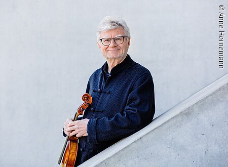 Ein älterer Mann steht hinter einer schräg verlaufenden, halbhohen Betonwand. Er stützt sich mit einem Arm darauf ab und hält eine Viola vor dem Oberkörper.