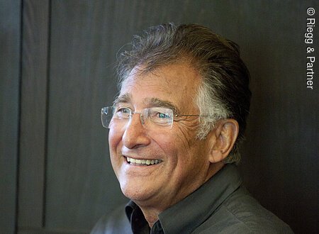 Prof. Siegfried Jerusalem Ein älterer Mann mit rahmenloser rechteckiger Brille sitzt vor einer Holzwand. Er schaut an der Kamera vorbei und lacht.