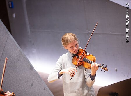 Violinspiel Hampe Altenburger Eine junge Frau im grauen Rollkragenpullover spielt Geige. Im Raumhintergrund sind geometrische Formen mit effektvoller Beleuchtung zu erkennen. Am linken Bildrand sind Teile einer Geige und eines Bogens zu sehen.