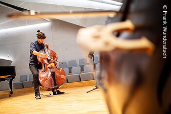Eine Person mit einem Kontrabass steht mit seinem Instrument in einem Raum mit Gesteinselementen. Er setzt den Bogen an..Am, rechten Bildrand ist unscharf der Steg eiees weiteren Kontrabasses zu sehen, links ein Teil eines Flügels.