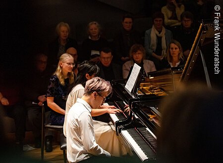 Klavierkonzert in Haus Marteau Eine junge Dame und ein Herr sitzen an einem Fl¸gel und spielt darauf. Im Hintergrund ist unscharf Publikum zu erkennen.