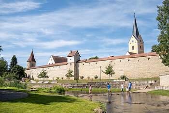 Familien mit Kindern überqueren Steg über Bach vor historischer Stadtmauer mit Kirchturm und blauem Himmel.